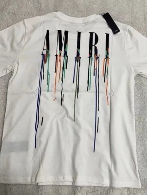 Vit AMIRI t-shirt med färgstänk - Säljer en vit t-shirt från AMIRI med stort tryck på ryggen där bokstäverna har färgstänk och droppar i blått, grönt, orange och svart. T-shirten har korta ärmar och en klassisk passform. Perfekt för dig som gillar streetwear och unika detaljer.