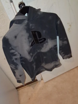 Grå PlayStation hoodie med batikmönster - Snygg grå hoodie med svart PlayStation-logga på bröstet och PS5-tryck på ryggen. Batikmönstrad design och stor känguruficka framtill. Perfekt för dig som älskar gaming och vill ha en chill stil. Mjuk och skön med huva.