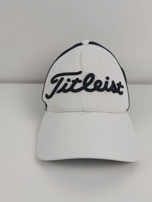 Titleist Pro V1 Tour Performance golf keps. Justerbar - Vit Titleist keps med svart mesh baktill. Kepsen har en justerbar snapback-stängning för en anpassad passform. Framsidan pryds av en broderad Titleist-logotyp i svart. På sidorna finns logotyper för FootJoy och Pro V1. Baktill finns en röd och vit Titleist-etikett. Finns lite smuts framme ( sista bild) annars i mycket bra skick!