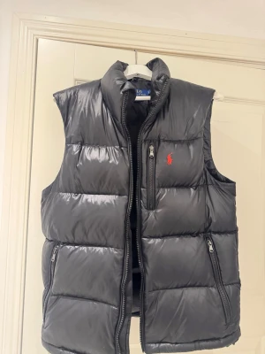 Svart dunväst från Polo Ralph Lauren - Snygg svart dunväst från Polo Ralph Lauren med klassisk röd logga på bröstet. Västen har hög krage, två sidofickor med dragkedja och en bröstficka med RL-dragkedja. Materialet är glansigt och quiltat för extra värme och stil. Använd max 5 ggr och inga skador. KAN DISKUTERA PRIS