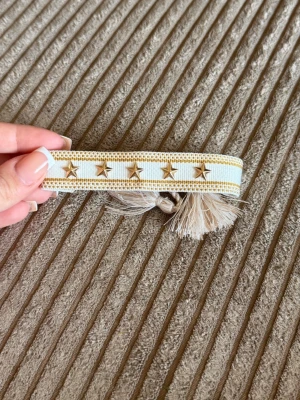 Beige tygarmband med guldstjärnor - Snyggt armband i beige och vitt tyg med dekorativa guldstjärnor och små nitar. Armbandet har fransiga kanter och knyts med tofsar. Perfekt accessoar för en trendig och avslappnad look.