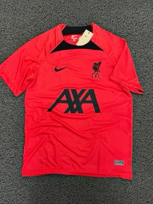 Liverpool tränings t-shirt röd Nike - Röd tränings t-shirt från Nike med Liverpool FC-logga och AXA sponsortryck i svart. T-shirten har korta ärmar, svart detalj vid halsen och är tillverkad i ett lätt, sportigt material som andas. Perfekt för fotbollsträning eller gym.