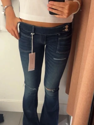 Mörkblå bootcut jeans med slitningar - Snygga mörkblå bootcut jeans med låg midja och slitningar vid knäna. Jeansen har dubbla knappar i midjan, dekorativa sömmar och fickor fram. Perfekta för en trendig och avslappnad look. Passar mig som är 160 cm.