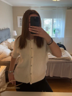 Vit fluffig kofta med pärlknappar - Supersöt vit kofta med korta ärmar och pärlknappar framtill. Koftan är fluffig och mjuk, perfekt att slänga över en topp för en chill look. Rak passform och enkel design som passar till det mesta.