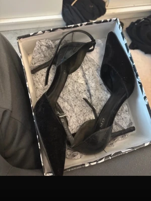 Svarta pumps med ankelrem från Tiamo - Säljer ett par snygga svarta pumps från Tiamo med spetsig tå och smal klack. Skorna har en elegant ankelrem med spänne och är tillverkade i sammet för en lyxig känsla. Perfekta för att lyfta din outfit.