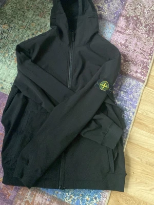 Stone Island svart vindjacka med huva - Svart vindjacka från Stone Island med huva och hel dragkedja. Jackan har den klassiska logotypen på vänster ärm och två fickor framtill. Materialet är lätt och smidigt, perfekt för dig som gillar streetwear och enkel stil.