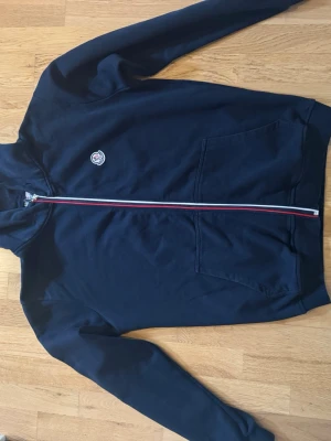 Mörkblå hoodie  - Snygg mörkblå hoodie från Moncler med dragkedja i rött, vitt och blått. Klassisk Moncler-logga på bröstet och två stora fickor framtill. Tillverkad i mjukt bomullsmaterial och har en huva för extra stilpoäng.