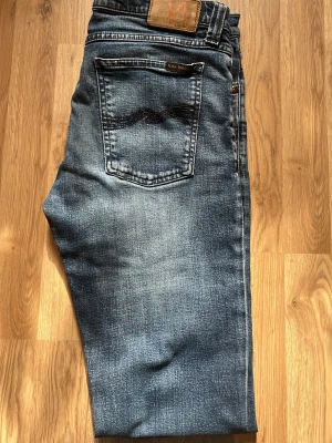 Blå jeans från Nudie Jeans - Snygga blå jeans från Nudie Jeans med klassisk femficksdesign och tydliga slitningar. Jeansen har en cool tvättad look, kontrastsömmar och läderpatch med logga bak i midjan. Materialet är denim i bomull.