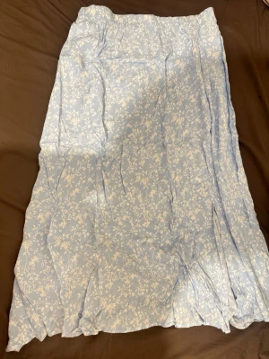 Ljusblå blommig midikjol - Söt ljusblå midikjol med vitt blommönster. Kjolen har en lätt utställd passform och är tillverkad i ett tunt, flowy tyg som ger en luftig känsla. Perfekt för dig som gillar romantiska och somriga vibes. Har även fickor och finns i både storlek L och M