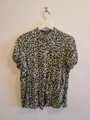Mönstrad kortärmad blus med knappar - Snygg blus med korta ärmar och knappar framtill. Mönstret är prickigt i svart, vitt, grått och grönt. Blusen har en liten ståkrage och är tillverkad i ett lätt, mjukt tyg som faller fint.
