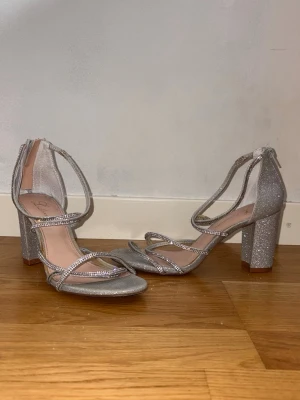 Glittriga silverpumps med blockklack - Snygga silverfärgade pumps med glittrigt material och smala remmar dekorerade med strass. Skorna har öppen tå och en stabil blockklack, samt dragkedja bak för enkel påtagning. Perfekta för att sticka ut och ge extra bling till din outfit.