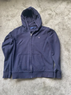 Mörkblå hoodie med dragkedja från Gant - Snygg mörkblå hoodie från Gant med dragkedja framtill och två stora fickor. Klassisk huva och diskret Gant-logga broderad på bröstet. Perfekt för en avslappnad och stilren look. Mjuk bomullskänsla och ribbade muddar vid ärmslut och nederkant.