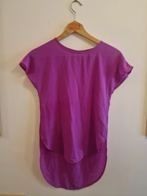 Lila topp från H&M med rund hals - Säljer en lila topp från H&M med rund halsringning och korta ärmar. Toppen har en längre bakdel för en cool och avslappnad look. Perfekt att styla med jeans eller kjol. Materialet känns lätt och luftigt.