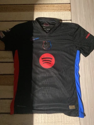 Svart FC Barcelona matchtröja Nike - Snygg svart FC Barcelona matchtröja från Nike med klubbmärke på bröstet, Spotify-logga i rött och blå/röda detaljer på sidorna. Tröjan har korta ärmar och sportig passform, perfekt för fotboll eller träning. Pris kan diskuteras 