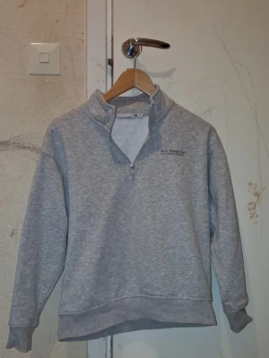 Grå halvzip hoodie Los Angeles (Man) - Snygg gråmelerad hoodie med halv dragkedja och hög krage. På bröstet står det 'Los Angeles' i svart text. Hoodien har ribbade muddar vid ärmar och nederkant. Perfekt för en chill och avslappnad stil.