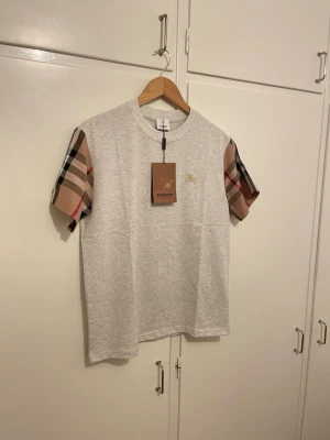 Grå Burberry t-shirt med rutiga ärmar - Säljer en grå t-shirt från Burberry med klassiska rutiga korta ärmar och broderad logga på bröstet. T-shirten har rund hals och är gjord i mjuk bomull. Perfekt för dig som vill ha en stilren look med exklusiva detaljer.