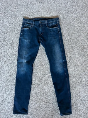 Blå anbass jeans från Replay - Säljer ett par blå slim fit jeans från Replay med snygga slitningar och klassisk femficksdesign. Jeansen har en mörk tvätt med ljusare partier och kontrastsömmar. Perfekta för dig som gillar en modern och stilren look.
