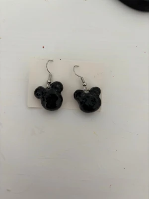 Svarta örhängen med musseform - Unika örhängen i svart med glansig finish, formade som Musse Pigg-huvuden. Hängande modell med krok i metall. Perfekta för dig som gillar lekfulla accessoarer och vill sticka ut lite extra.