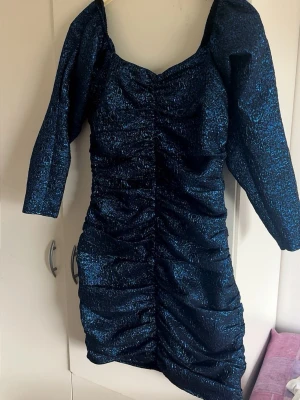 Glittrig blå festklänning - Snygg glittrig blå kortklänning. Klänningen har en figurnära passform som framhäver siluetten. 