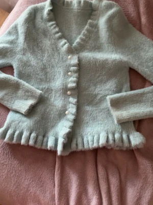 Ljusblå fluffig kofta med volang - Står Strl L men passar S-M Supersöt ljusblå kofta med volangkant och pärlknappar. Koftan är fluffig och har v-ringning samt långa ärmar. Perfekt för dig som gillar en mjuk och feminin stil. Materialet känns mysigt och koftan har en figurnära passform.