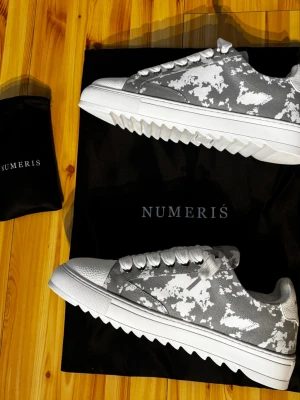 Gråvita sneakers från Numeris - Snygga sneakers från Numeris i grått och vitt camomönster. Skorna har kraftig sula med taggig design, vita skosnören och tåparti i textil. Märkeslogga på plösen. Perfekta för dig som vill sticka ut med en unik look.