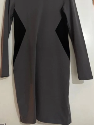 Grå långärmad klänning H&M - Snygg grå långärmad klänning från H&M med svarta triangulära paneler i sidorna som ger en cool kontrast. Klänningen har en rak passform och rund halsringning. Perfekt för dig som gillar minimalistisk och stilren design.