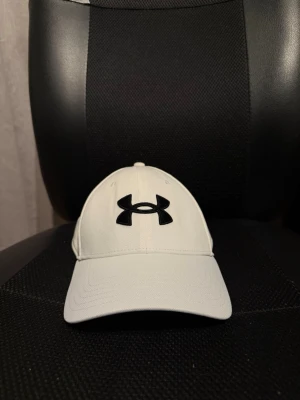 Vit keps från Under Armour - Snygg vit keps från Under Armour med klassisk böjd skärm och svart logga framtill. Kepsen har broderad text bak och är tillverkad i ett mjukt och slitstarkt material som passar perfekt till sportiga outfits.