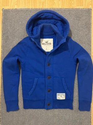 Vintage Hollister Hoodiejacka - Säljer denna feta Hollister hoodiejackan, fodrad inne så varm och skön till det kalla vädret | Väldigt bra skick förutom lite tecken på användning se sista bild | Storlek S | Längd ≈ 65cm, Bredd armhåla - armhåla ≈ 52cm | Modell 175cm, 68kg