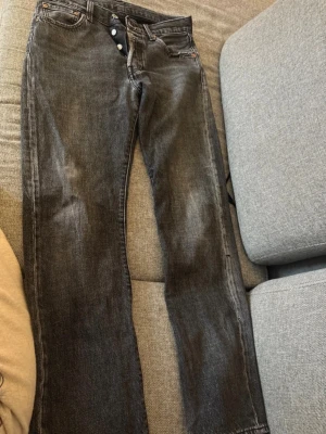 Svarta raka jeans med knappgylf - Säljer ett par svarta jeans med raka ben och knappgylf. Jeansen har en klassisk femficksmodell och är tillverkade i denim. Färgen är tvättad svart med en något sliten look. Perfekta för en avslappnad och trendig stil. Se skavanker på bilderna. 