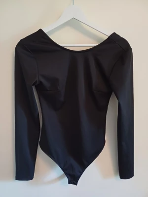 Svart långärmad body - Snygg svart body med långa ärmar och djup rygg. Tillverkad i ett stretchigt, slätt material som sitter tight mot kroppen. Perfekt för att skapa en clean och stilren look. Skriv om ni har frågor, priset går att diskutera ❤️