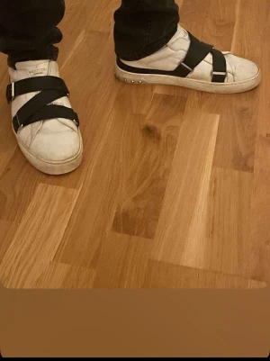 Valentino vita sneakers med svarta band - Säljer ett par vita sneakers från Valentino med breda svarta band över foten och silverdetaljer på sulan. Skorna har en låg profil och en chunky sula som ger en cool kontrast. Perfekta för dig som vill sticka ut med en unik design. Skorna är lite använda men kan tvättas vid köp kontakta mig om du har några vidare frågor. Skorna kostar i vanliga fall 5k 