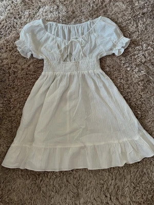 Vit kort omlottklänning med volang - Söt vit kortklänning med puffärmar, omlottdetalj och volangkant nedtill. Klänningen har smock i midjan för en bekväm passform och är tillverkad i ett lätt, luftigt tyg som passar perfekt till varma dagar.