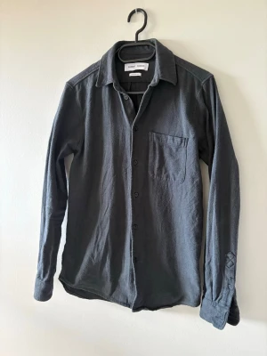 Mörkgrå skjorta från Samsoe Samsoe - Stilren mörkgrå skjorta från Samsoe Samsoe med klassisk krage, bröstficka och knäppning framtill. Skjortan har långa ärmar och är gjord i ett mjukt, strukturerat tyg som ger en avslappnad men snygg look.