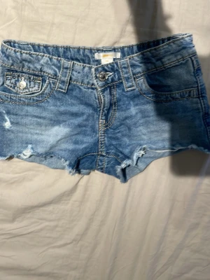 Blå jeansshorts med rå kant - Snygga blå jeansshorts med råa kanter och slitningar. Klassisk femficksmodell med knapp och dragkedja framtill samt bälteshällor. Perfekta för varma dagar och ger en avslappnad vibe.