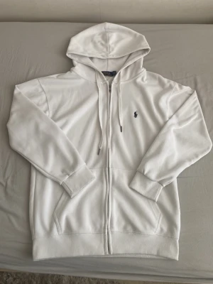 Vit hoodie från Polo Ralph Lauren  - Säljer en stilren vit hoodie från Polo Ralph Lauren med dragkedja framtill och klassisk huva med dragsko. Tröjan har två fickor fram och den ikoniska lilla marinblå loggan broderad på bröstet. Perfekt för en clean och avslappnad look.