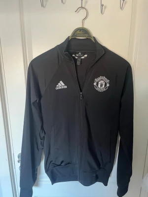 Manchester United full zip  - Svart full zip från Adidas med Manchester United-logga på bröstet. Jackan har hel dragkedja, ståkrage och vita broderade detaljer. Perfekt för fotbollsträning eller som supporterplagg.