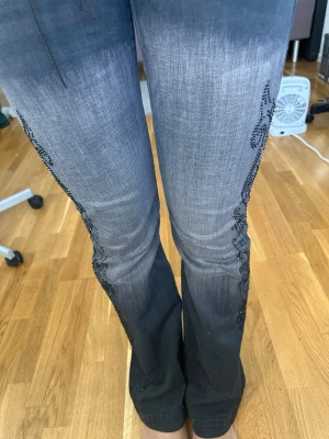 Grå bootcut jeans med snörning - Säljer ett par grå bootcut jeans med svart snörning i midjan och dekorativa svarta pärlor längs sidorna. Jeansen har en snygg tvättad look och utsvängda ben. Perfekta för dig som vill sticka ut lite extra!