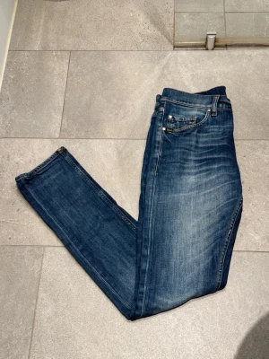 Blå raka jeans med slitningar - Säljer ett par klassiska blå jeans med raka ben och snygga slitningar på framsidan. Jeansen har fem fickor, normal midja och är tillverkade i denim. Perfekta för en avslappnad och trendig look. Jeansen är i nyskick och endast använda en gång, säljer eftersom de inte sitter bra på mig, Mitt pris 449, nypris 1600kr, storleken är 28/34 men på tiger of sweden jeans får man räkna bort ungefär 2 i längd så dessa sitter ungefär 28/32