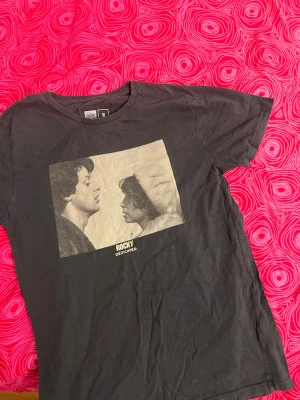 Svart Rocky t-shirt från Dedicated -  Svart t-shirt från Dedicated med ett stort tryck av en klassisk Rocky-film scen. 