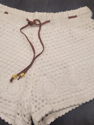 Vita virkade shorts med snörning från Ibiza. - Säljer ett par vita virkade shorts med brun flätad snörning och guldiga detaljer. Shortsen har ett genombrutet mönster och dekorativa cirklar längs kanten. Perfekta för sommaren och ger en chill boho-vibe.