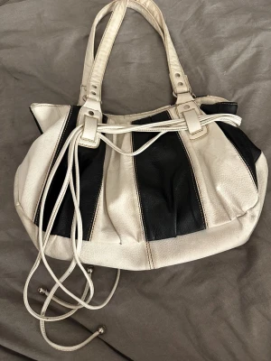 Randig svartvit handväska - Snygg handväska i svart och vitt med breda ränder och långa axelband. Väskan har dekorativa snören framtill och silverfärgade detaljer. 