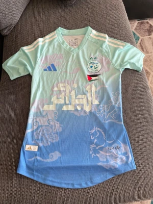 Ljusblå fotbollströja från Adidas - Snygg ljusblå fotbollströja från Adidas med vita och blå detaljer, klubbmärke och flagga på bröstet samt arabiskt tryck framtill. Tröjan har korta ärmar, V-ringning och HEAT.RDY-material som andas. Perfekt för träning eller match.