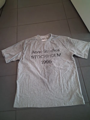 Grå Acne Studios t-shirt - Snygg grå t-shirt från Acne Studios med trycket 'Acne Studios STOCKHOLM 1996' på bröstet. Klassisk passform med rund hals och korta ärmar. Perfekt basplagg för en clean och avslappnad stil.