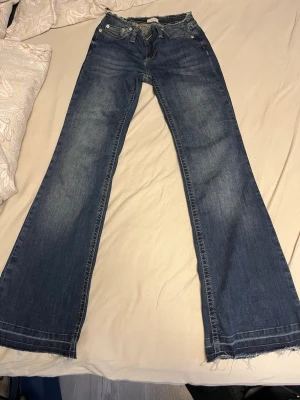 Blå bootcut jeans från Nelly  - Säljer ett par blå bootcut jeans från nelly med råa kanter i midjan och vid bensluten. Jeansen har klassisk femficksdesign, slitningar och ljusare partier framtill för en cool vintagekänsla. Perfekta för dig som gillar en avslappnad och trendig stil.