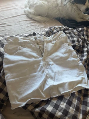 Vit jeanskjol med rå kant - Säljer en vit kort jeanskjol med rå, fransig kant nertill. Kjolen har klassisk femficksdesign, bälteshällor och dragkedja framtill. Perfekt till sommaren för en fräsch och avslappnad look.