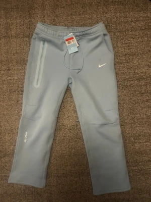 Nike NOCTA ljusblå mjukisbyxor - Snygga ljusblå mjukisbyxor från Nike NOCTA med raka ben, dragsko i midjan och dragkedjeförsedd ficka på sidan. Byxorna har diskreta loggor och är tillverkade i ett mjukt, stretchigt material som känns riktigt skönt mot huden.