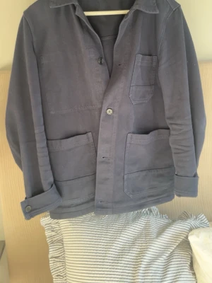 Blå overshirt jacka från arket  - Stilren blå overshirt jacka från arket med tre stora fickor framtill och knäppning med knappar. Jackan har klassisk krage och raka ärmar, perfekt för lager på lager. Tillverkad i ett mjukt bomullsmaterial som känns bekvämt och avslappnat.