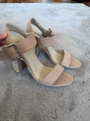 Beige rosa klackar med bred rem - Snygga beige rosa klackar med bred rem över foten och justerbart spänne i silver. Skorna har en chunky klack och öppen tå. Materialet är mockaimitation som ger en mjuk och stilren look. 