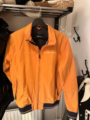 Orange vindjacka Sail Racing - Orange vindjacka från Sail Racing med dragkedja framtill, två sidofickor med dragkedja och logga på bröstet. Jackan har ribbade muddar med ränder i svart, vitt och rött samt en liten logga i nacken. Perfekt för blåsiga dagar.