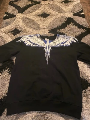 Svart sweatshirt med vingeprint Marcelo Burlon - Cool svart sweatshirt från Marcelo Burlon med ett stort vitt och blått vingeprint runt halsen. Tröjan har rund hals och ribbade muddar. Perfekt för dig som vill sticka ut med en unik design från County of Milan. Passar L